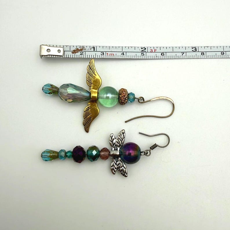 May include: Two dangle earrings featuring angel-shaped charms with beads. The top earring has a gold-tone wing charm, a green bead, and a faceted iridescent bead. The bottom earring has a silver-tone wing charm and multi-coloured beads. A ruler shows a length of approximately 7.6 cm.