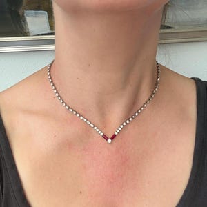 May include: A silver-toned necklace featuring a V-shaped center point. The chain is composed of small, round, clear stones, while the focal point at the bottom consists of two rectangular red stones flanking a single round clear stone at the apex.