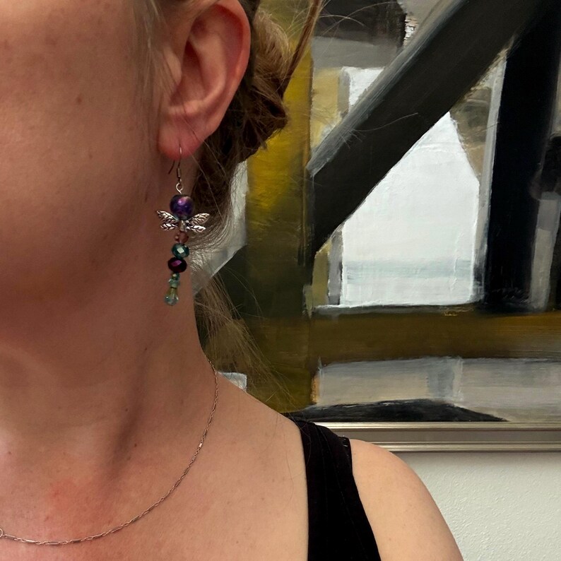 May include: A dangle earring featuring a silver-toned dragonfly charm positioned between a large, iridescent purple bead at the top and a series of smaller, faceted beads in shades of dark purple, teal, and light green hanging vertically below.