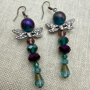 May include: A pair of drop earrings featuring silver-toned metal dragonfly charms. Each earring is composed of a large iridescent purple bead at the top, followed by a vertical arrangement of faceted glass beads in shades of teal, green, and purple.