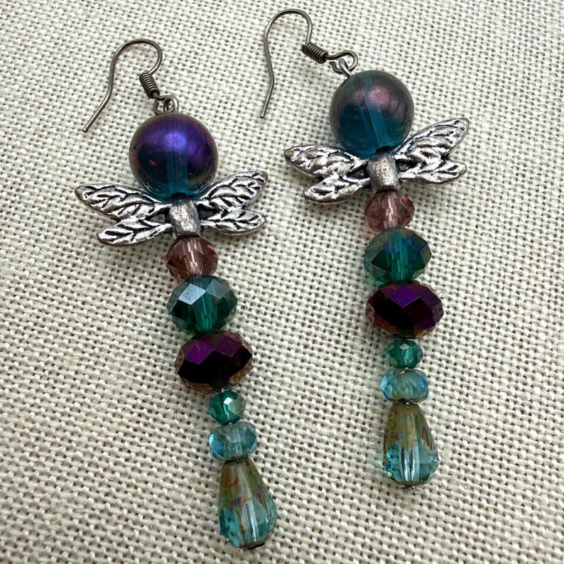May include: A pair of drop earrings featuring silver-toned metal dragonfly charms. Each earring is composed of a large iridescent purple bead at the top, followed by a vertical arrangement of faceted glass beads in shades of teal, green, and purple.