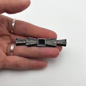 May include: A black bow-shaped brooch with a square center and rhinestone accents. The brooch has a rectangular shape and is held in a hand against a white background. The bow design is a classic accessory.