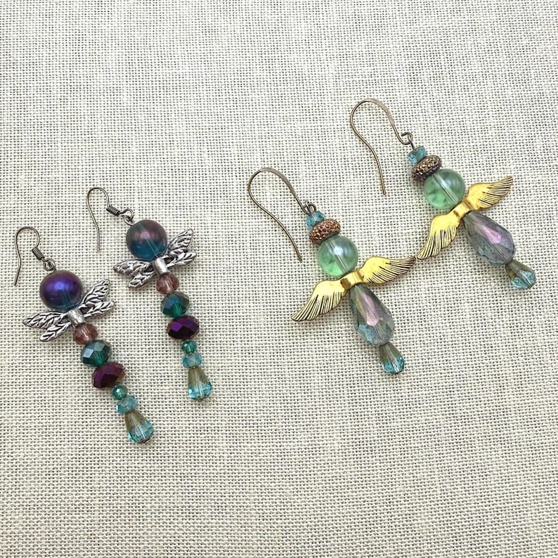 May include: Two pairs of dangle earrings featuring angel-shaped designs. The left pair has silver-toned metal wings with purple and teal beads. The right pair has gold-toned metal wings with light green and iridescent teardrop-shaped glass beads.