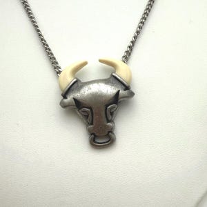 May include: A silver-toned bull pendant necklace with a cream-colored horn design. The bull's head features a ring at the bottom and is suspended from a silver chain. The necklace is displayed against a white background.