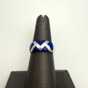 May include: A silver band ring featuring a repeating geometric pattern of inlaid deep blue stone segments. The metal surface displays a polished finish, and the design consists of alternating triangular and chevron-shaped blue stone inlays.