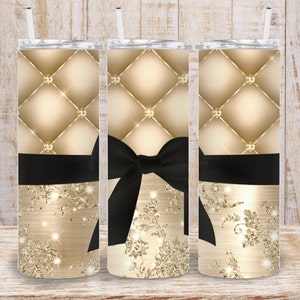 Könnte beinhalten: Drei goldene und schwarze Edelstahlbecher mit einem gesteppten Design, einer schwarzen Schleife und goldenen Glitzerakzenten.