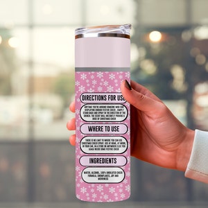 May include: A pink and silver insulated tumbler with a lid. The tumbler has a snowflake pattern and text that reads "Directions for Use", "Where to Use", and "Ingredients". The text describes how to use the Christmas cheer spray.