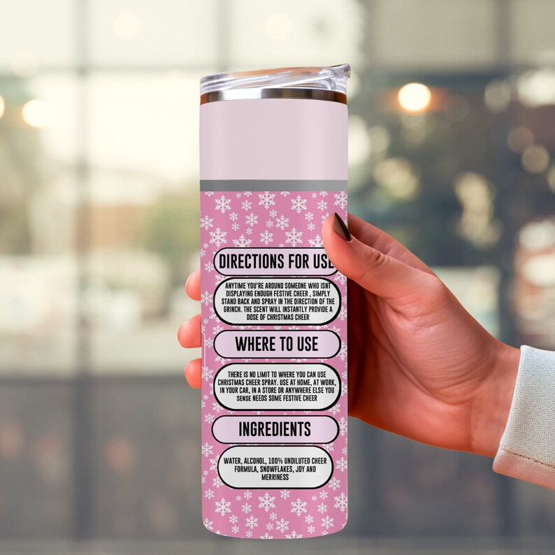 May include: A pink and silver insulated tumbler with a lid. The tumbler has a snowflake pattern and text that reads "Directions for Use", "Where to Use", and "Ingredients". The text describes how to use the Christmas cheer spray.