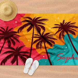 May include: A colourful beach towel with a sunset scene featuring palm trees and the name "Sophia" printed on it. The towel is laid out on a sandy beach with a straw hat and flip flops.