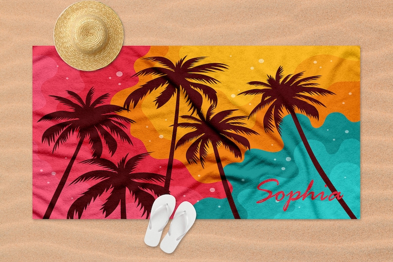 May include: A colourful beach towel with a sunset scene featuring palm trees and the name "Sophia" printed on it. The towel is laid out on a sandy beach with a straw hat and flip flops.