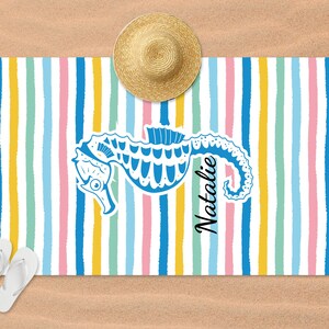 Puede incluir: Una toalla de playa colorida con un patr&oacute;n a rayas en rosa, amarillo, azul y verde. Un caballito de mar azul est&aacute; en el centro de la toalla con el nombre "Natalie" escrito verticalmente a su lado.