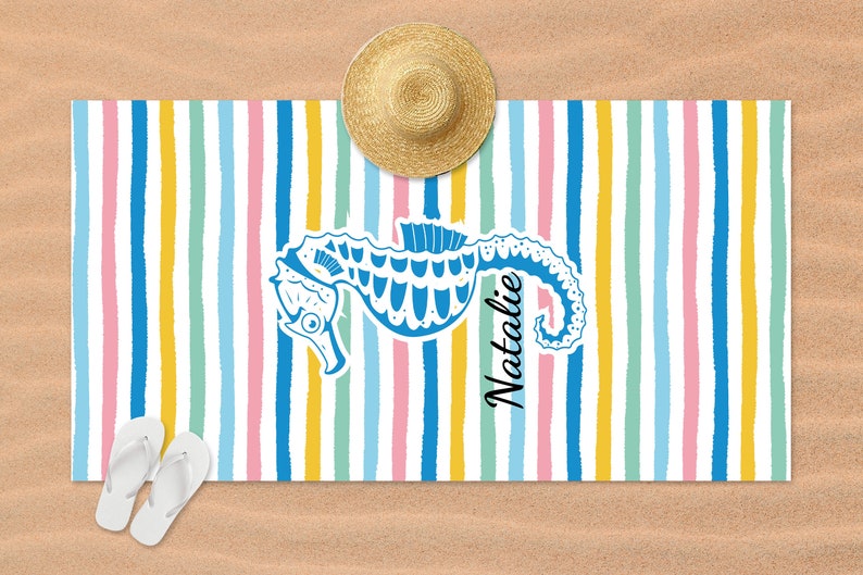 Puede incluir: Una toalla de playa colorida con un patr&oacute;n a rayas en rosa, amarillo, azul y verde. Un caballito de mar azul est&aacute; en el centro de la toalla con el nombre "Natalie" escrito verticalmente a su lado.
