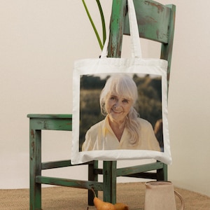 May include: A white tote bag with a photo of a woman with gray hair smiling. The bag is hanging on a green wooden chair. There are pears in a brown bowl and a beige pitcher on a woven rug.