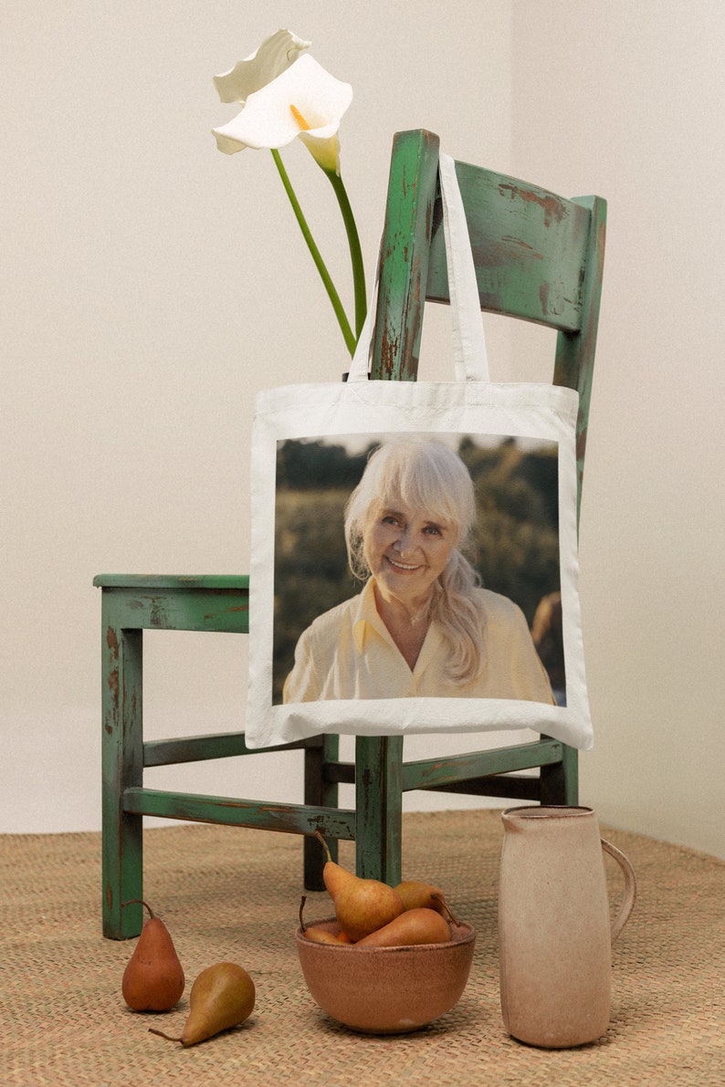 May include: A white tote bag with a photo of a woman with gray hair smiling. The bag is hanging on a green wooden chair. There are pears in a brown bowl and a beige pitcher on a woven rug.