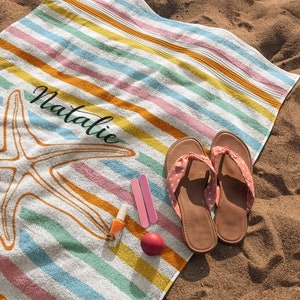 Puede incluir: Una toalla de playa a rayas de colores con un dise&ntilde;o de estrella de mar y el nombre "Natalie" impreso en ella. La toalla est&aacute; sobre una playa de arena con un par de chanclas rosas y esmalte de u&ntilde;as.