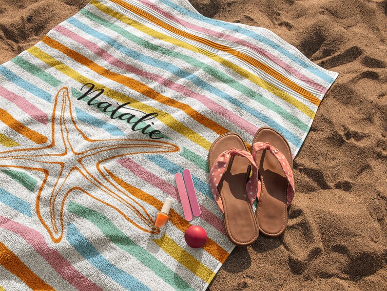 Puede incluir: Una toalla de playa a rayas de colores con un dise&ntilde;o de estrella de mar y el nombre "Natalie" impreso en ella. La toalla est&aacute; sobre una playa de arena con un par de chanclas rosas y esmalte de u&ntilde;as.