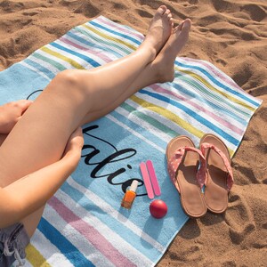 Puede incluir: Una toalla de playa a rayas con la palabra "Vacation" escrita en negro. La toalla est&aacute; en una playa de arena con una persona sentada en ella. La persona lleva sandalias y tiene esmalte de u&ntilde;as en los dedos de los pies.