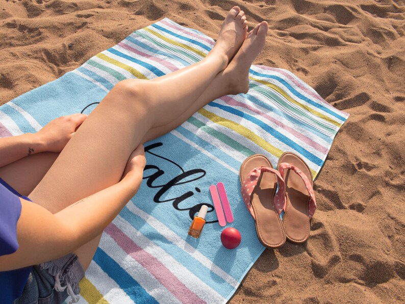 Puede incluir: Una toalla de playa a rayas con la palabra "Vacation" escrita en negro. La toalla est&aacute; en una playa de arena con una persona sentada en ella. La persona lleva sandalias y tiene esmalte de u&ntilde;as en los dedos de los pies.