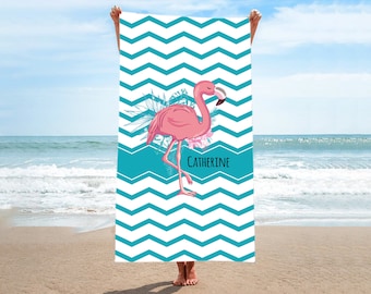 May include: A white beach towel with a turquoise chevron pattern. A pink flamingo with black eyes and a black beak stands on the towel. The towel has the name "Catherine" printed on it.