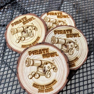 May include: Four wooden coasters with a rustic, natural edge. Each coaster features a laser-etched design of a cannon with the words "PIRATE BOWL" and "MASTER GUNNER 300 GAME". The coasters are made of light brown wood.
