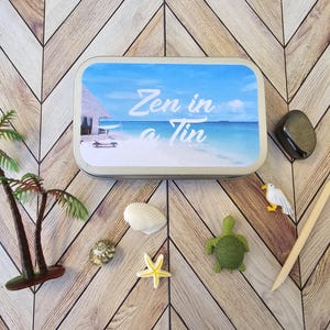 May include: A silver tin with a blue and white beach scene and the text "Zen in a Tin". The tin is surrounded by various small decorative items, including a seashell, a starfish, a turtle, and a seagull.