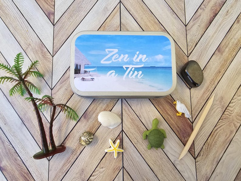 May include: A silver tin with a blue and white beach scene and the text "Zen in a Tin". The tin is surrounded by various small decorative items, including a seashell, a starfish, a turtle, and a seagull.