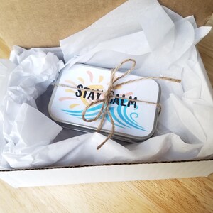 May include: A small, rectangular tin with the words "STAY CALM" printed on it, tied with twine, rests inside a white box with tissue paper. The tin features a sun and wave design in shades of yellow and blue.