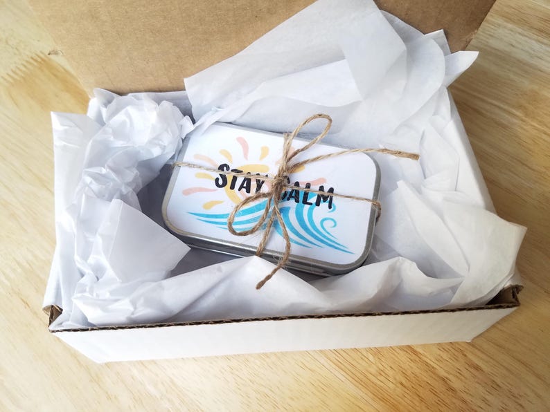 May include: A small, rectangular tin with the words "STAY CALM" printed on it, tied with twine, rests inside a white box with tissue paper. The tin features a sun and wave design in shades of yellow and blue.