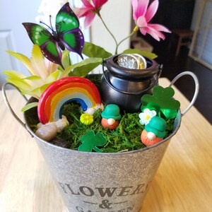 May include: A small metal bucket with the words "FLOWER & GARDEN" printed on the side. The bucket is filled with green moss, a rainbow, two gnomes, a pot of gold, and a butterfly.