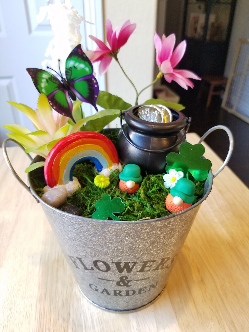 May include: A small metal bucket with the words "FLOWER & GARDEN" printed on the side. The bucket is filled with green moss, a rainbow, two gnomes, a pot of gold, and a butterfly.