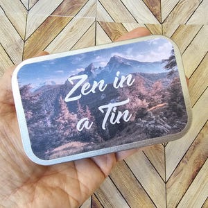 Pu&ograve; includere: Una scatola di metallo rettangolare con un design di paesaggio montano. L'immagine presenta una catena montuosa, alberi e il testo "Zen in a Tin" in scrittura bianca. La scatola &egrave; tenuta in mano.