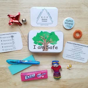 Könnte beinhalten: Eine Sammlung von Gegenständen zur Angstlinderung, darunter eine Metalldose mit dem Text "I am Safe" und einer Baumgrafik, eine Windmühle, ein Stift, ein Stressring und Karten mit Bewältigungsaussagen. Ebenfalls enthalten ist eine Packung Extra-Kaugummi.
