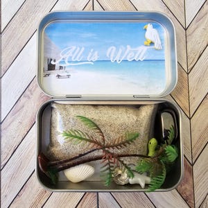 May include: A metal tin with a beach scene on the lid. The lid says "All is Well". The tin is open and contains sand, a small palm tree, a seashell, a small seagull, and a green snake.