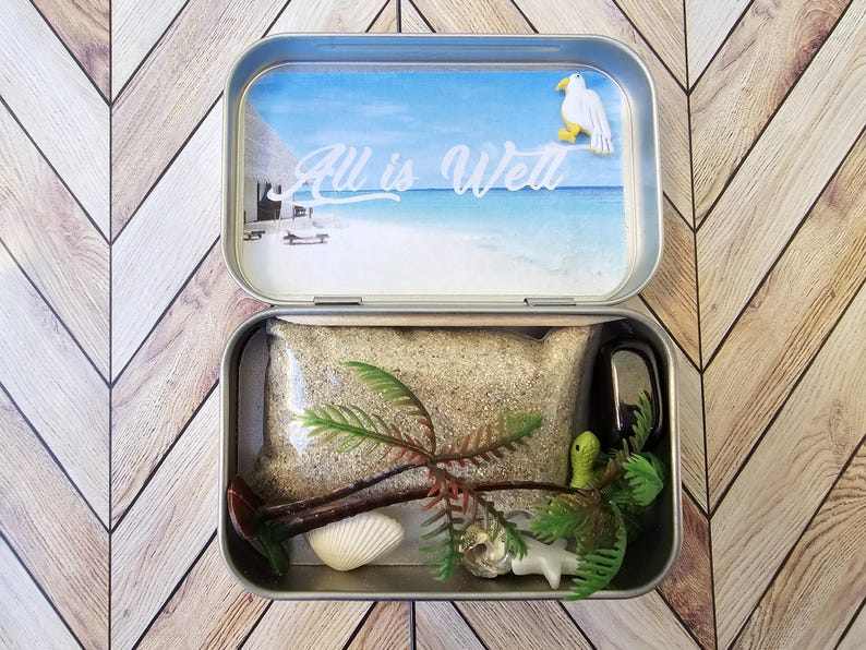 May include: A metal tin with a beach scene on the lid. The lid says "All is Well". The tin is open and contains sand, a small palm tree, a seashell, a small seagull, and a green snake.