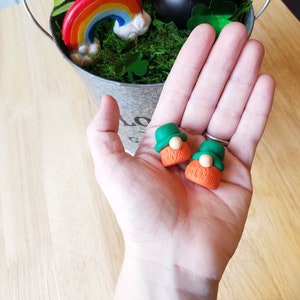May include: Two small green and orange gnome figurines with white faces and beards. The gnomes are sitting on a hand.