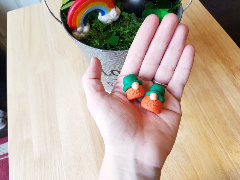 May include: Two small green and orange gnome figurines with white faces and beards. The gnomes are sitting on a hand.
