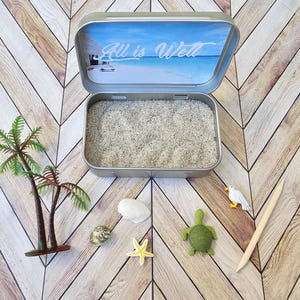 May include: A small metal tin with a blue and white beach scene on the lid. The lid is open and the tin is filled with white sand. There are small plastic palm trees, a sea turtle, a seagull, a starfish, and a seashell on the table around the tin. The lid of the tin has the text "All is Well" printed on it.