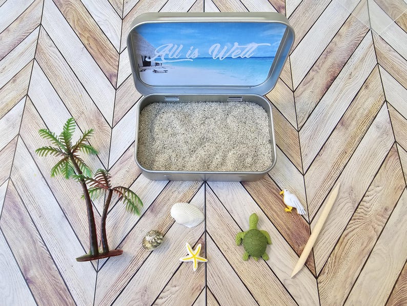 May include: A small metal tin with a blue and white beach scene on the lid. The lid is open and the tin is filled with white sand. There are small plastic palm trees, a sea turtle, a seagull, a starfish, and a seashell on the table around the tin. The lid of the tin has the text "All is Well" printed on it.