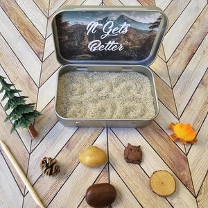 Pu&ograve; includere: Una scatola d'argento aperta con le parole "It Gets Better" e una scena di montagna all'interno, riempita di sabbia. Un piccolo albero verde, un bastoncino di legno, una pigna e altri piccoli oggetti decorativi sono disposti intorno alla scatola.