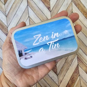 May include: A silver tin with a blue and white beach scene and the text "Zen in a Tin" printed on the front.
