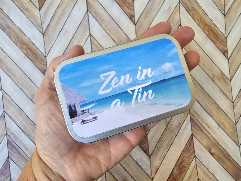 May include: A silver tin with a blue and white beach scene and the text "Zen in a Tin" printed on the front.