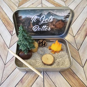 Pu&ograve; includere: Una scena di campeggio in miniatura all'interno di una scatola di latta argentata. La scena comprende un piccolo albero verde, una pietra marrone, una pigna, una fetta di legno e un fal&ograve;. Il coperchio presenta la scritta "It Gets Better".