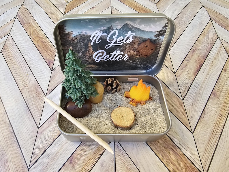 Pu&ograve; includere: Una scena di campeggio in miniatura all'interno di una scatola di latta argentata. La scena comprende un piccolo albero verde, una pietra marrone, una pigna, una fetta di legno e un fal&ograve;. Il coperchio presenta la scritta "It Gets Better".