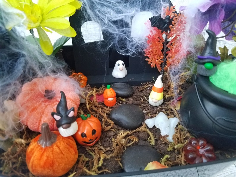 Op de afbeelding: Een Halloween-thema miniatuur feeentuin met een zwart huis, een geest, een dwerg, een ketel, pompoenen en spinnenwebben.