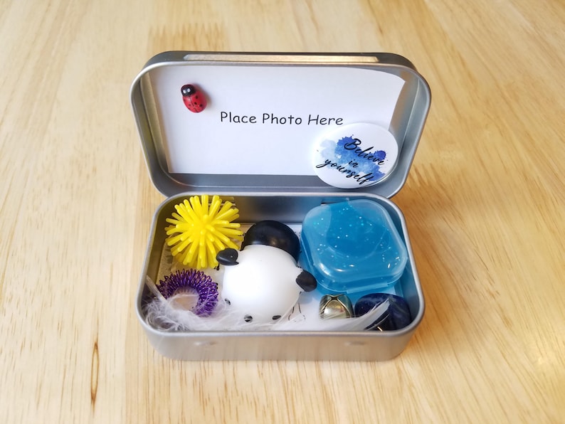 May include: A small, open metal tin filled with various items. Inside are a yellow spiky ball, a purple ring, a white snowman figure, a blue gel-like item, and other small objects. The lid has a ladybug and a sticker that says "Believe in yourself."