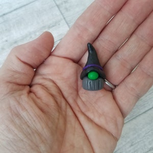 Op de afbeelding: Een klein heksengnomfiguurtje gemaakt van polymer clay. De gnom draagt een zwarte punthoed met een paarse band en heeft een groen oog. De baard van de gnom is gemaakt van grijze klei en heeft een houtnerfstructuur.