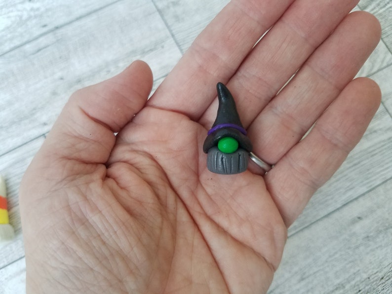 Op de afbeelding: Een klein heksengnomfiguurtje gemaakt van polymer clay. De gnom draagt een zwarte punthoed met een paarse band en heeft een groen oog. De baard van de gnom is gemaakt van grijze klei en heeft een houtnerfstructuur.