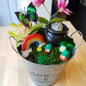May include: A small metal bucket with a green shamrock design and the words "Flower & Garden" on the side. The bucket is filled with moss, a rainbow, a pot of gold, and two leprechaun figurines.