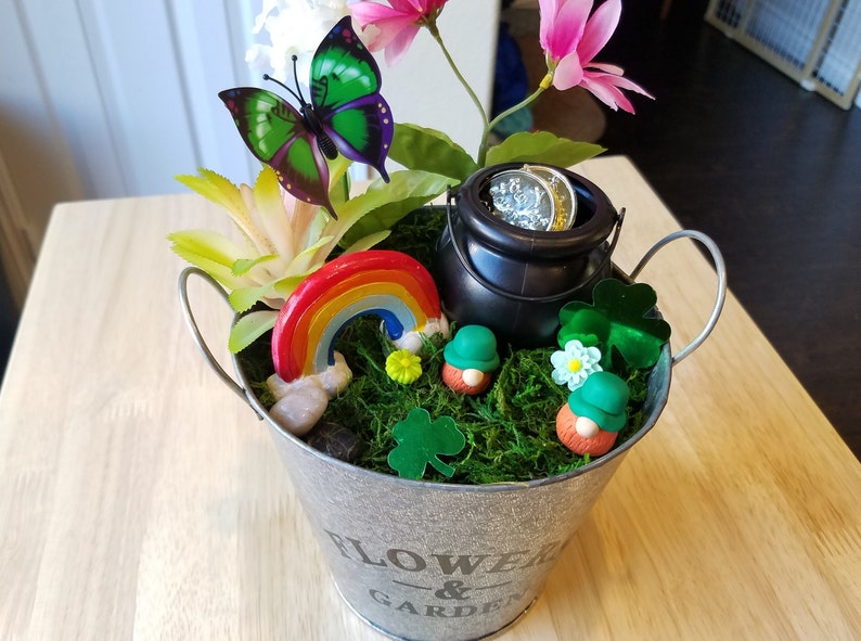 May include: A small metal bucket with a green shamrock design and the words "Flower & Garden" on the side. The bucket is filled with moss, a rainbow, a pot of gold, and two leprechaun figurines.