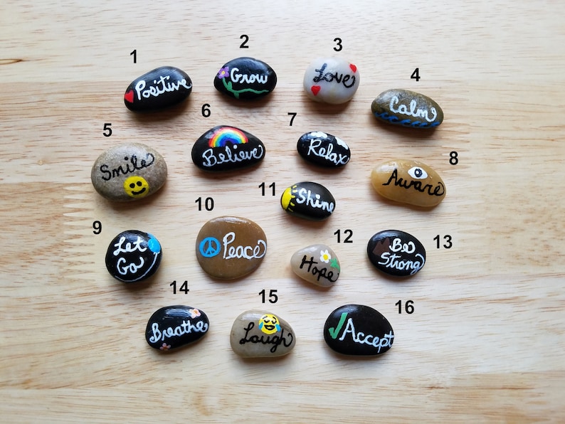 Op de afbeelding: Een set van 16 beschilderde stenen met positieve affirmaties. De stenen zijn in verschillende vormen en maten en zijn geschilderd in zwart, wit, bruin, geel, blauw, groen en rood. De affirmaties zijn: "Positief", "Groeien", "Liefde", "Kalm", "Glimlach", "Geloven", "Ontspannen", "Bewust", "Loslaten", "Vrede", "Stralen", "Hoop", "Sterk zijn", "Ademhalen", "Lachen" en "Accepteren".